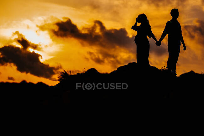 Low angle side view silhouettes of unrecognizable couple expecting baby standing back to back and holding hands while admiring sundown in mountains together — Stock Photo