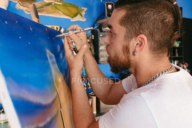 Vista lateral do artista masculino usando pistola de pulverização para pintar quadro em tela durante o trabalho em oficina criativa — Fotografia de Stock