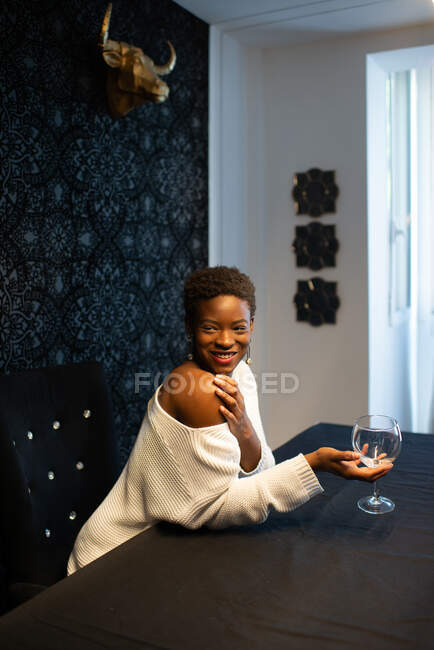 Side view of cheerful African American female with glass of refreshing cocktail sitting at table and chilling in dark room during weekend — Stock Photo