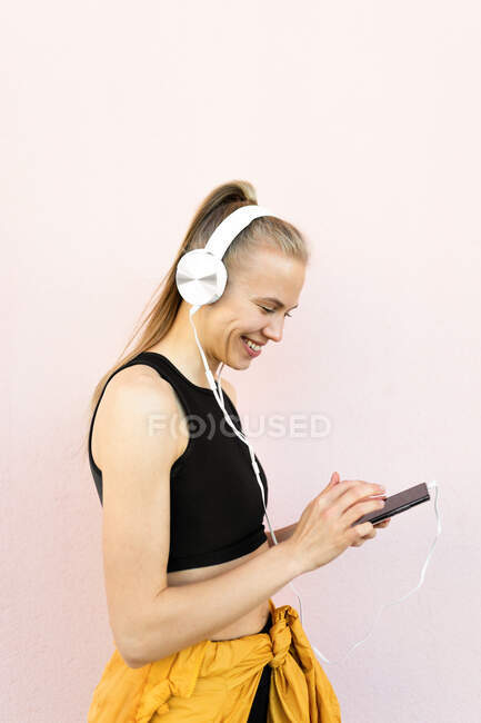 Junge kaukasische Frau trägt Kopfhörer und Sportkleidung, hört Musik am Telefon und lächelt, isoliert auf hellem Hintergrund — Stockfoto