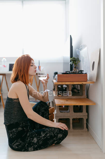 Side view of young female with glass of alcoholic drink listening to song from vintage record player at home — Stock Photo