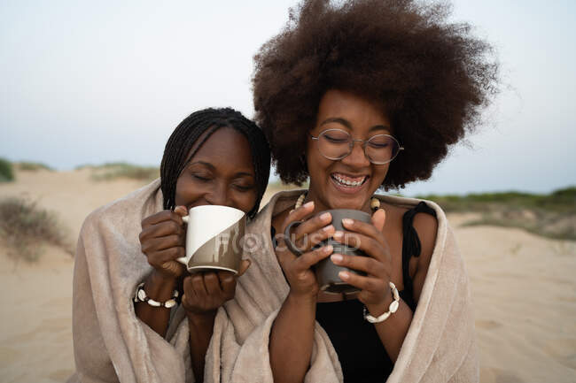 Счастливые молодые чернокожие подруги с кружками горячих напитков сидят рядом, завернутые в теплое одеяло, и веселятся летним вечером на песчаном пляже — стоковое фото
