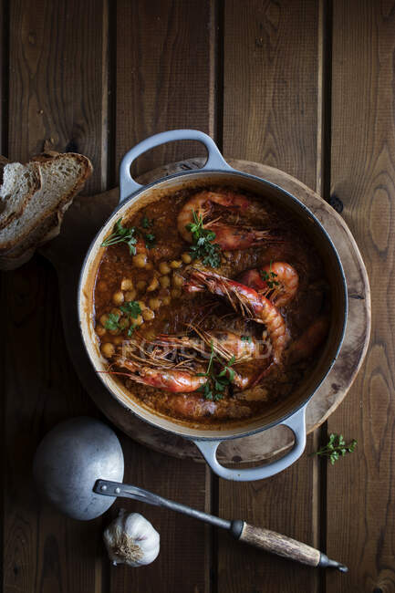 Top view of white saucepan with delicious homemade stew with chickpeas and shrimps placed on round wooden tray next to rustic metal plate with ladle and garlic and some croutons on wooden background — Stock Photo