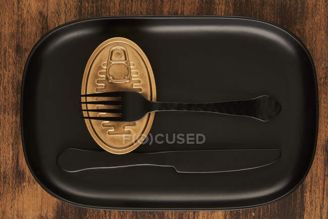 Верхний вид черной вилки и ножа помещен рядом с запечатанной консервной пищей на прямоугольном черном подносе — стоковое фото