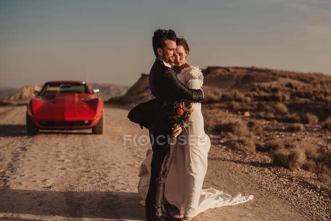 Side view of bride and groom embracing each other while standing near red luxury vehicle during road trip through Bardenas Reales Natural Park in Navarra, Spain — Stock Photo