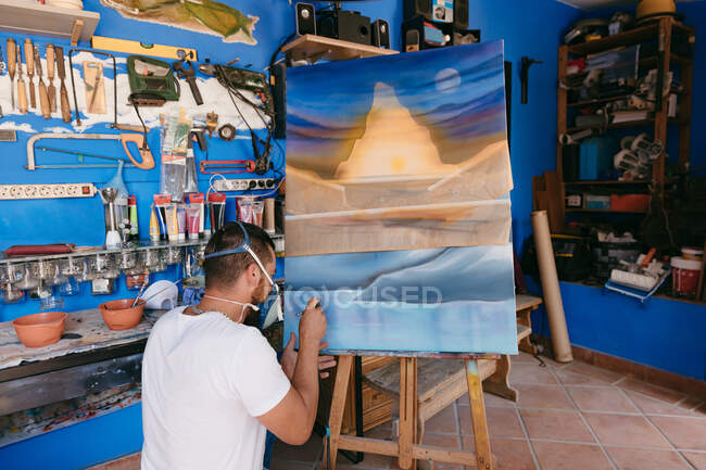 Man in respirator spraying paint on canvas with abstract landscape while working in professional creative studio — Stock Photo