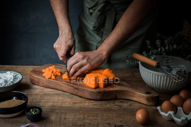 Неузнаваемый мужчина-повар в фартуке рубит сырую тыкву на деревянной разделочной доске рядом с мукой и хлебными крошками с семенами и яйцами во время приготовления пирога — стоковое фото