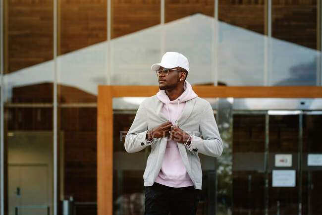 Confident African American male wearing casual clothes and cap and sunglasses standing against modern building and looking away — Stock Photo