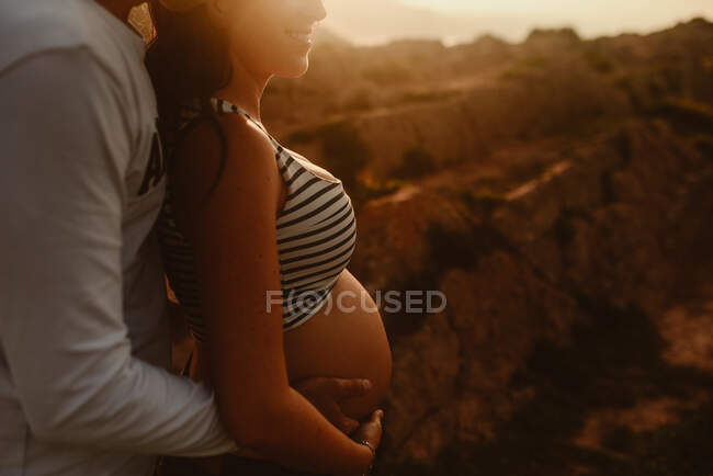 Vista lateral del hombre irreconocible abrazando a la feliz esposa embarazada mientras están de pie juntos contra el paisaje montañoso y admirando la puesta de sol en la noche de verano - foto de stock
