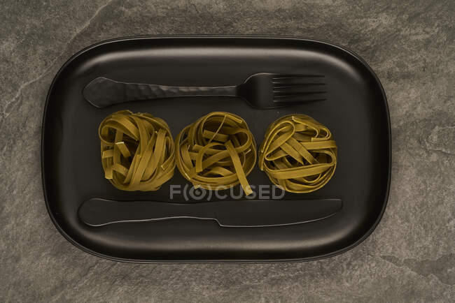 Top view of black fork and knife placed near uncooked tagliatelle pasta on tray on table — Stock Photo