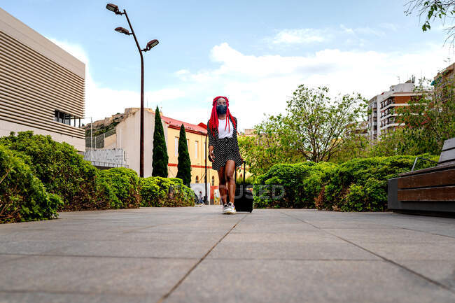 Молодая афроамериканка в защитной маске с яркими дредами прогуливается в парке с чемоданом и смотрит в сторону — стоковое фото