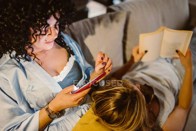 Schnittwinkel Frau schaut Handy mit lesbischem Partner lesen Buch in der Freizeit zu Hause — Stockfoto