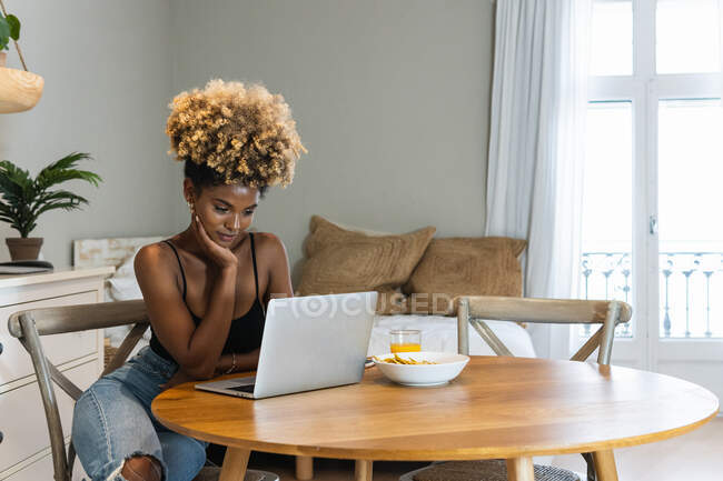 Привлекательная афроамериканка-фрилансер сидит на стуле и смотрит за ноутбуком за завтраком — стоковое фото