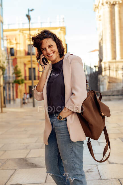 Adulto ispanico femminile in tendenza casual strappato jeans e blazer con zaino sopra la spalla ridendo felicemente durante la conversazione telefonica mentre in piedi sulla piazza urbana — Foto stock