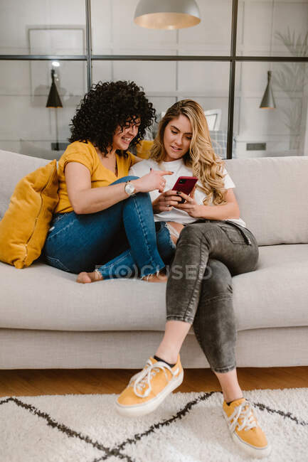 Все тело веселой лесбийской пары в повседневной одежде просматривая мобильный телефон и сидя вместе на удобном диване в современной гостиной — стоковое фото