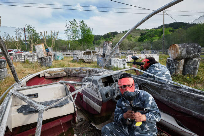 Unrecognizable men with guns and in protective helmets playing paintball among abandoned boat and cars — Stock Photo