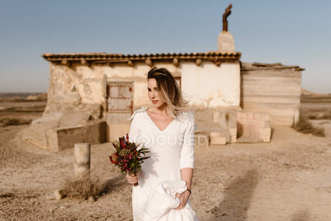 Donna in abito bianco e con fiori in piedi contro cabina invecchiata squallida il giorno del matrimonio nel Parco Naturale di Bardenas Reales in Navarra, Spagna — Foto stock
