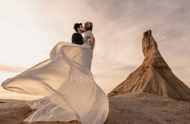 Angolo basso di sposo e sposa che si baciano vicino alla montagna contro il cielo nuvoloso al tramonto nel Parco Naturale di Bardenas Reales in Navarra, Spagna — Foto stock