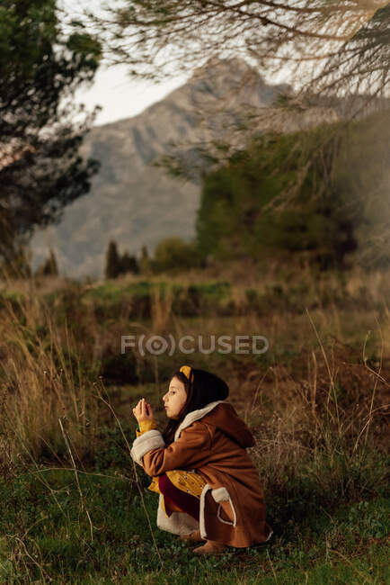 Vista laterale di una bambina curiosa in abiti caldi che osserva l'ambiente mentre trascorre del tempo da sola sul prato erboso contro le montagne — Foto stock
