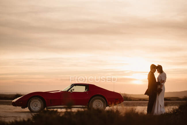 Мужчина и женщина в элегантной одежде касаются лба, стоя возле спортивного автомобиля на фоне закатного неба во время свадебного торжества в природном парке Барденас-Реалес в Наварре, Испания — стоковое фото