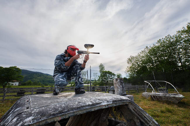 Male player in camouflage outfit and with gun sitting on abandoned car and aiming while playing paintball in countryside — Stock Photo
