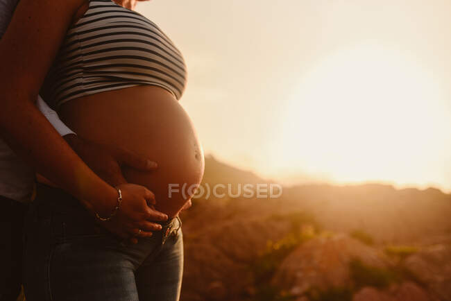 Вид збоку на невпізнаваного чоловіка, який приймає щасливу вагітну дружину, стоячи разом проти гірського пейзажу та захоплюючого заходу сонця в літній вечір — стокове фото