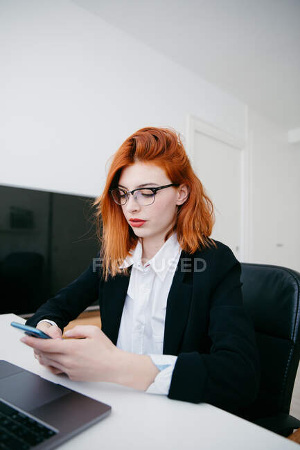 Молода жінка-підприємець у формальному носінні текстових повідомлень на мобільний телефон під час роботи за столом з ноутбуком вдома — стокове фото