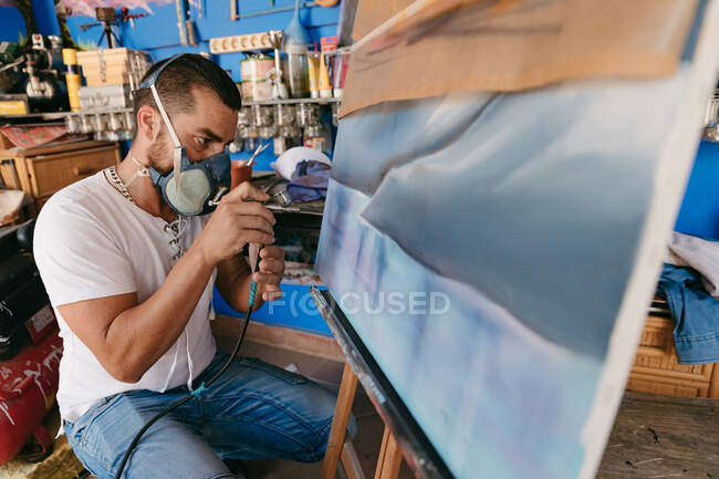 Side view of male artist in respirator using spray gun to paint picture on canvas during work in creative workshop — Stock Photo