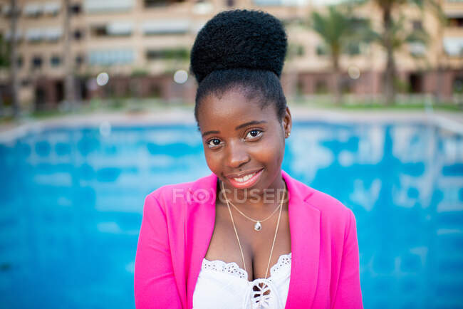 Jeune femme afro-américaine élégante avec un chignon afro-américain portant un blazer rose vif et un haut blanc avec des accessoires souriants à la caméra contre la piscine extérieure floue — Photo de stock