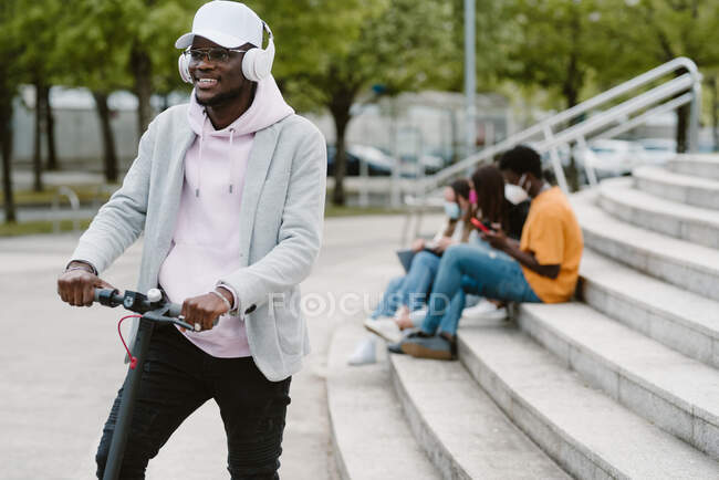 Hombre afroamericano en auriculares inalámbricos montando scooter eléctrico con amigos irreconocibles sentados en la escalera en el fondo - foto de stock