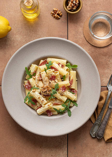 Dall'alto piatto di pasta con rucola, salame e noci su un tavolo circondato da olio, limone e posate — Foto stock