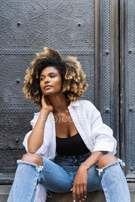 Confiada mujer afroamericana con elegante atuendo casual sentada y mirando hacia otro lado en la escalera de hormigón contra puertas de metal - foto de stock