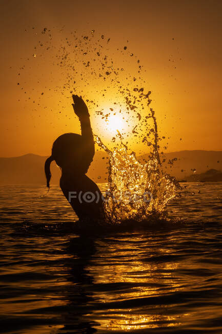 Vista laterale silhouette di anonima bambina in piedi in acqua di mare e fare schizzi con le mani contro la luce del tramonto in estate — Foto stock