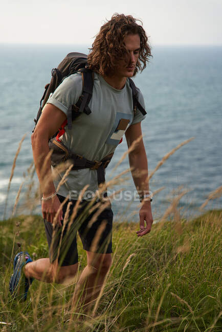 Mochilero masculino caminando en la colina durante el trekking en verano y mirando hacia abajo - foto de stock