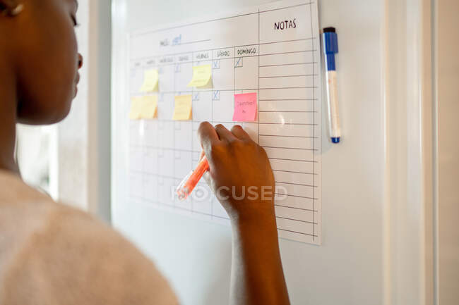 Side view of crop ethnic female writing in paper planner hanging on refrigerator in kitchen — Stock Photo