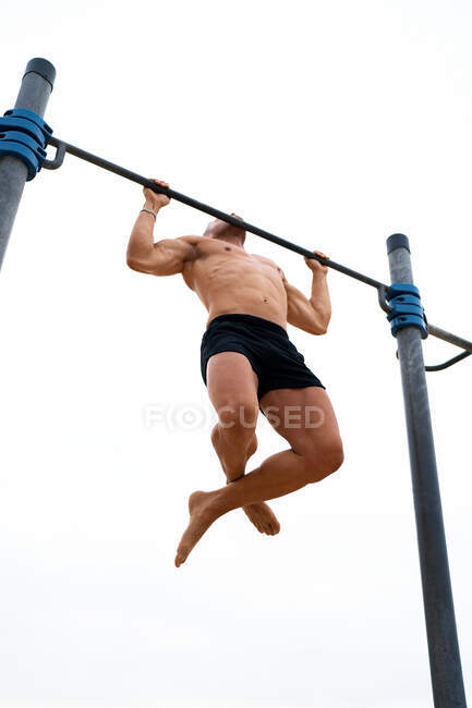 Low angle of muscular male athlete with naked torso doing chin ups on horizontal bar during training on background of gray sky — Stock Photo