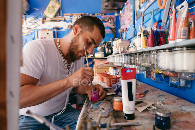 Homem barbudo em roupas casuais mexendo pigmento em jarra com pincel enquanto sentado à mesa em oficina criativa — Fotografia de Stock