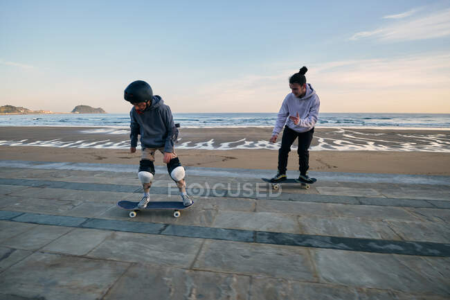 Молоді активні друзі чоловічої статі катаються на скейтбордах вздовж набережної на фоні моря і заходу сонця — стокове фото