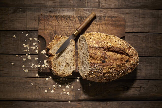 Верхний вид вкусного свежеиспеченного хлеба из запеченного теста с нарезанным куском и ножом, помещенным на разделочную доску на столе — стоковое фото