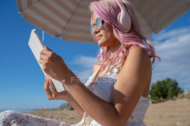 Vista lateral de ángulo bajo de la mujer con el pelo rosa escalofriante en la orilla del mar mientras escucha música en los auriculares y navegar por Internet en la tableta en verano - foto de stock