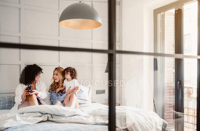 Счастливая молодая лесбийская пара и симпатичный малыш отдыхают на уютной кровати в современной светлой спальне — стоковое фото