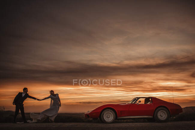 Braut und Bräutigam halten Händchen neben rotem Sportwagen vor wolkenverhangenem Sonnenuntergang im Bardenas Reales Naturpark in Navarra, Spanien — Stockfoto
