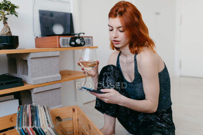 Вид на молодую женщину со стаканом алкогольного напитка, слушающую песню из винтажного проигрывателя и просматривающую телефон — стоковое фото