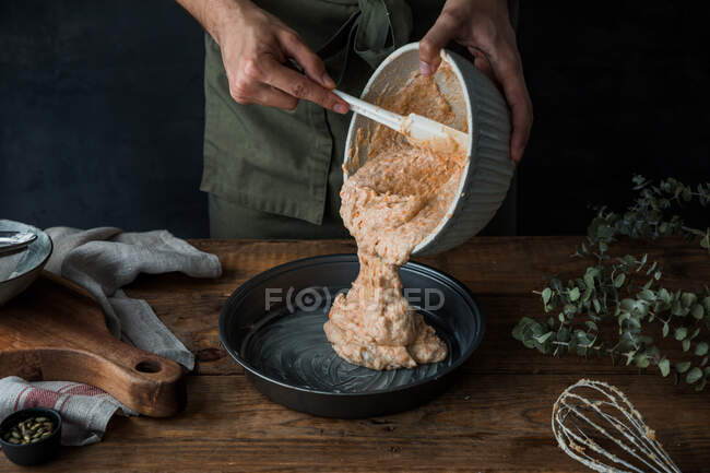 Anonymous man in apron using silicone spatula to pour from a bowl batter into a tin while preparing pumpkin pie on lumber table — Stock Photo