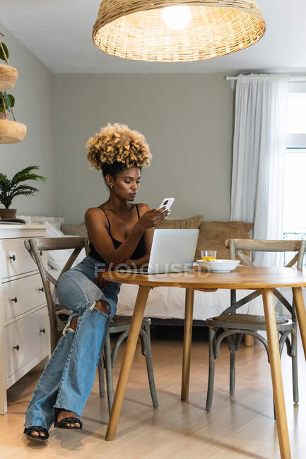 Full body of African American female freelancer sitting on chair and watching laptop while browsing on smartphone at table with breakfast — Stock Photo