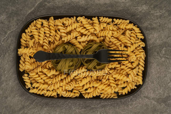 Vista superior do garfo preto colocado perto de massa fusilli não cozida na bandeja na mesa — Fotografia de Stock