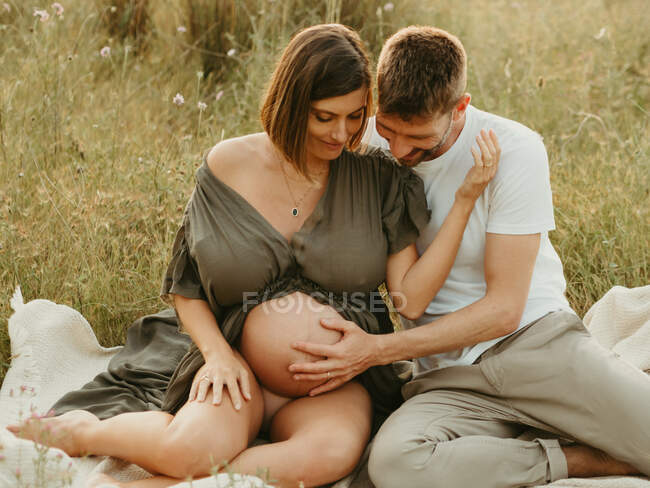 Приємні обійми вагітної жінки з закритими очима, коли сидять на лузі в сільській місцевості. — стокове фото
