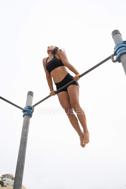 Dal basso dell'atleta donna in forma che fa esercizio muscolare sulla barra durante l'allenamento contro il cielo grigio — Foto stock