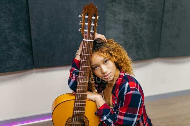 Giovane chitarrista femminile etnica con i capelli ricci appoggiati alla chitarra acustica mentre guarda la fotocamera — Foto stock