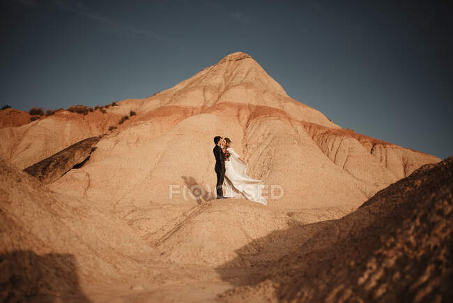 Vista laterale dello sposo e della sposa che si abbracciano guardandosi vicino alle montagne contro il cielo nuvoloso al tramonto nel Parco Naturale di Bardenas Reales in Navarra, Spagna — Foto stock